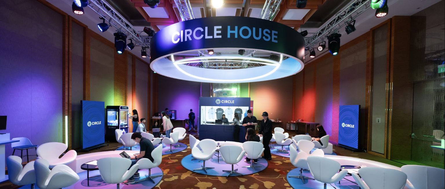 The Circle House lounge at TOKEN2049, a lit overhead CIRCLE HOUSE ring above white lounge seating and colour-washed walls