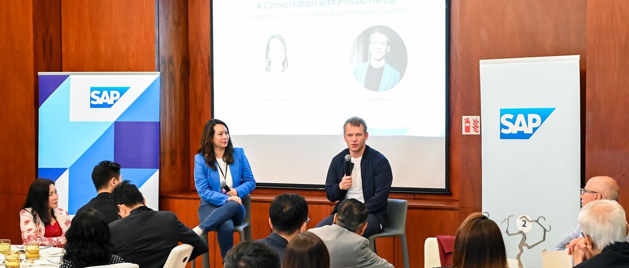 A fireside conversation between two speakers seated between SAP banners, guests watching from the room's round tables