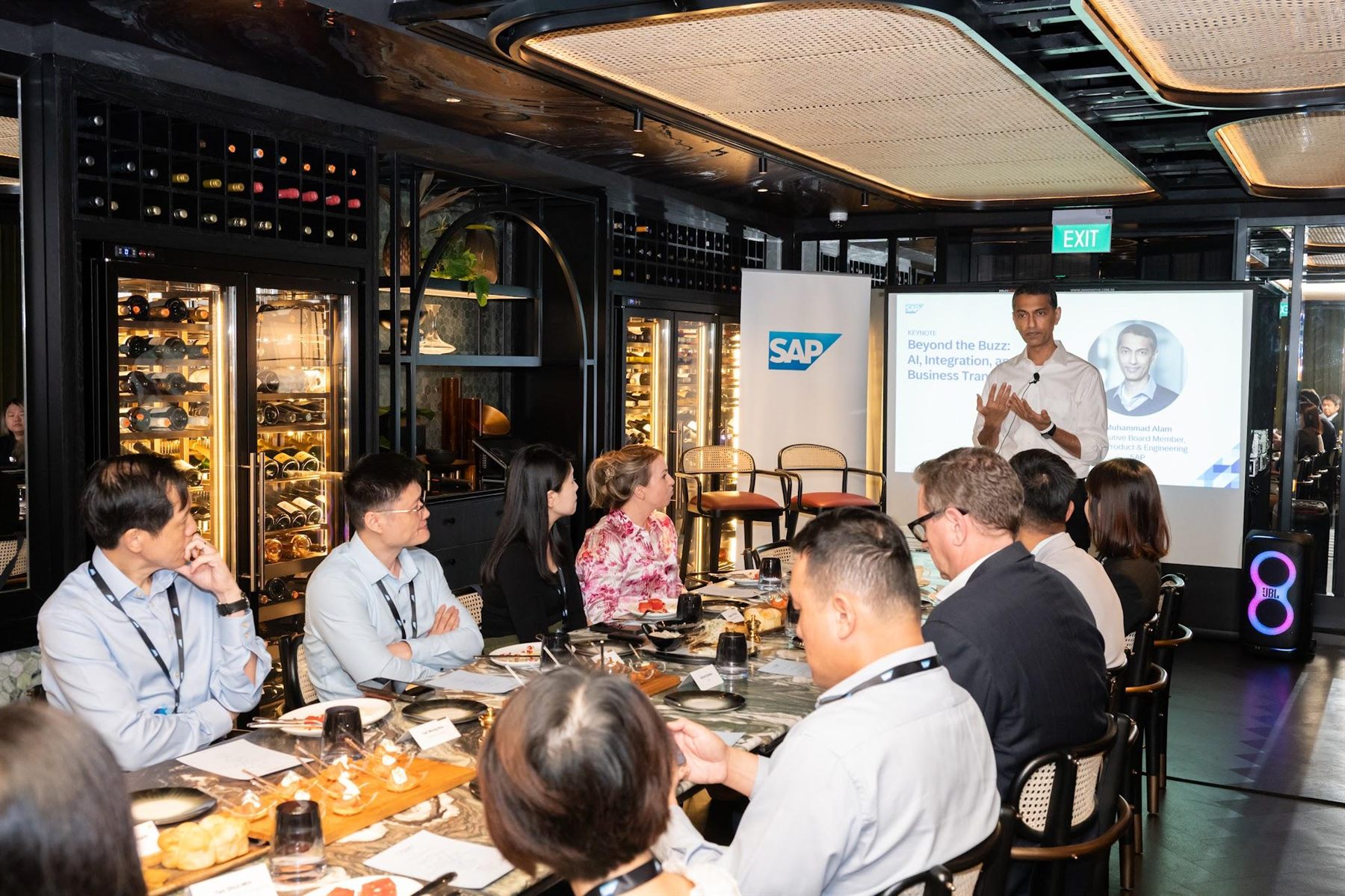 A private-dining keynote in a wine-cellar setting, guests seated along a marble table facing the speaker